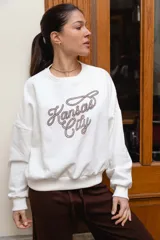 Buzo de felpa blanco con cuello redondo, mangas largas y puños elastizados, con estampa frontal en color marrón que dice "Kansas City" en tipografía cursiva con efecto de soga.