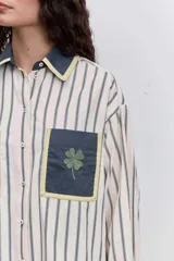 Camisa de corte oversize con estampado de rayas verticales, cuello clásico y cierre frontal de botones. Presenta dos bolsillos frontales en contraste con bordados de tréboles, además de puños y cuello en color sólido con detalles en amarillo.