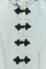 Chaleco de denim claro con efecto lavado, cuello mao, sin mangas y corte peplum con pliegues en la parte inferior. Cierre frontal con botones de alamares negros.