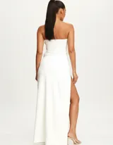 Vestido largo de fiesta color blanco, con escote strapless y detalles de pedrería en el borde superior. Presenta un diseño entallado con drapeado lateral y una abertura profunda en la pierna con detalle de pedrería en la unión.