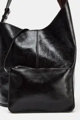 Bolso de hombro tipo saca asimétrica, color negro, con acabado de piel brillante. Posee un asa de hombro ajustable y cierre magnético. Incluye una cartera interior extraíble.