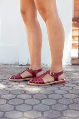 Sandalias planas estilo romano tipo cangrejera, confeccionadas en cuero charolado de color bordó. Presentan diseño de tiras entrelazadas en el empeine, puntera cuadrada, talón cerrado con tira ajustable mediante hebilla metálica dorada y suela plana con pequeño tacón.