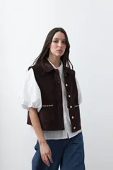 Chaleco de pana color marrón oscuro, con cuello camisero, cierre frontal de botones metálicos dorados y dos bolsillos frontales con detalle de bordado geométrico.