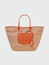 Bolso tipo tote de gran tamaño confeccionado en tejido de paja con rayas multicolores. Presenta un parche frontal de cuero naranja con el logo de la marca grabado, asas de hombro de cuero a juego y un detalle colgante de cuentas de madera. Incluye un cierre de mosquetón metálico en la base.
