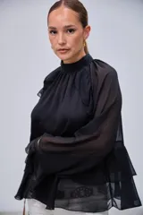 Blusa negra de cuello alto con volados en el pecho y mangas largas con transparencias. Presenta un diseño escalonado con volados en la parte inferior.