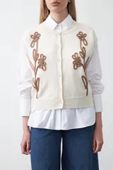 Chaleco tejido color crema con cuello redondo y cierre frontal de botones. Presenta un diseño bordado en relieve de color marrón con motivos florales y orgánicos en el frente.