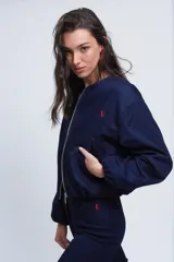 Campera tipo bomber de gabardina de jean color azul oscuro, con cuello a la base, cierre frontal metálico y bolsillos ojal. Presenta mangas largas abullonadas con puños elásticos y costuras con pespuntes en color rojo.