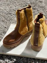 Botas de cuero marrón con la caña recubierta de corderito verde. Presentan una suela de plataforma tipo track en color beige y un tirador en la parte posterior para facilitar el calce.