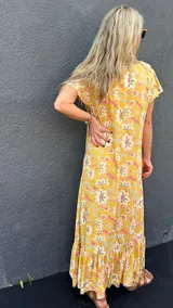 Vestido largo amarillo con estampado floral en tonos rosa, blanco y celeste. Tiene mangas cortas con volados y un volante en el ruedo.