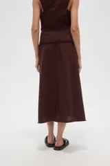 Falda larga de algodón color marrón oscuro, de corte envolvente (wrap skirt) con abertura lateral y borde deshilachado. Se ajusta con un lazo lateral que pasa por argollas metálicas.