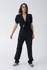 Mono negro de lino con escote pronunciado en V y nudo frontal que deja ver un corte en la cintura. Mangas cortas abullonadas y pantalón de corte recto.