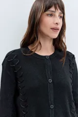 Cardigan negro de punto con acabado brillante, cuello redondo, cierre frontal de botones y detalle de cordones laterales entrelazados. Presenta mangas tres cuartos con diseño acampanado.
