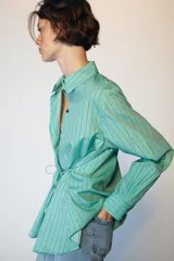 Camisa de manga larga con cuello solapa, cierre frontal con botones y diseño cruzado con lazada lateral en el mismo tejido. Presenta un estampado de rayas verticales en tonos verdes y azules.