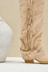 Bota texana alta de gamuza color beige, con taco cuadrado, flecos largos colgantes en el lateral y tiras con hebilla metálica alrededor del tobillo.