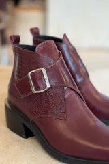 Botin de cuero color bordó, con diseño de puntera cuadrada y detalle de hebilla metálica lateral. Presenta una textura combinada en el lateral y suela de goma negra.