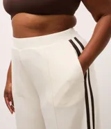 Pantalón deportivo de corte oversized estilo college, confeccionado en microfibra color crema con rayas laterales contrastantes en color marrón. Presenta cintura fija, bolsillos funcionales y frisos frontales.