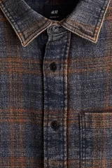 Camisa de franela de algodón suave con efecto lavado, corte relajado. Presenta cuello inglés, tapeta clásica de botones, un bolsillo superior abierto en el pecho y canesú en la espalda. Las mangas son largas con tapeta y puños abotonados, y el bajo es ligeramente redondeado. El estampado es a cuadros en tonos grises y naranjas.