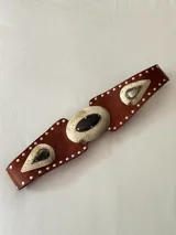 Cinto tipo faja de cuero color chocolate, con apliques metálicos plateados grabados y piedras incrustadas. Presenta detalles de tachas pequeñas en todo el borde y un diseño central ovalado.