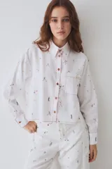 Camisa de corte recto y largo cropped, confeccionada en tela blanca con estampado de figuras deportivas pequeñas. Presenta cuello clásico, cierre frontal con botones de madera y costuras en hilo rojo a contraste. Incluye un bolsillo frontal en el pecho.