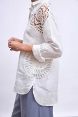 Camisa blanca de lino con cuello clásico, cierre frontal con botones y mangas largas con puños abotonados. Presenta apliques de encaje en hombros y bolsillos delanteros.