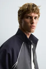 Cazadora relaxed fit en color block, con cuerpo gris oscuro y mangas azul marino. Presenta cuello subido, manga larga, bolsillos frontales de vivo, vivos combinados a contraste en blanco, acabados con rib elástico y cierre frontal con cremallera.