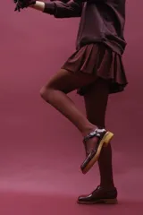 Zapato tipo guillermina de cuero charol color marrón oscuro, con tira T y hebilla ajustable en el lateral. Posee puntera cuadrada y suela de goma color caramelo con taco bajo.