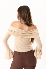 Blusa beige con hombros descubiertos, confeccionada en tela elástica con destellos de lurex. Presenta mangas largas con terminación acampanada y diseño ajustado al cuerpo.