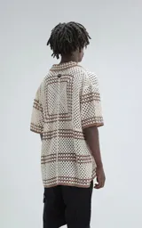 Camisa de manga corta tejida en punto abierto (símil crochet), color beige con detalles en marrón. Presenta cuello solapa, cierre frontal con botones y calce oversize.