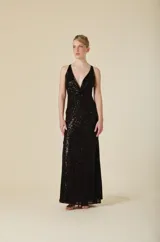 Vestido largo de fiesta color negro, completamente cubierto de lentejuelas. Presenta un escote en V profundo, tirantes anchos y un detalle de nudo en la cintura que genera un efecto drapeado, realzando la silueta.