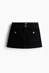 Pollera mini de denim negro con tiro bajo, cierre frontal y dos bolsillos delanteros tipo cargo. Presenta botones decorativos con detalles de strass en la cintura y en las solapas de los bolsillos.