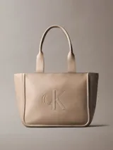 Cartera tote Calvin Klein color beige con asas dobles de mano y cierre de cremallera superior. Presenta el monograma de la marca grabado en el frente.