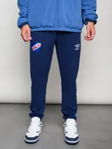 Pantalón de jogging azul marino con puños ajustados en los tobillos. Presenta un parche bordado del escudo del Club Nacional de Football en la pierna izquierda y el logo de Umbro en la derecha. Tiene detalles en azul claro en la parte inferior de las piernas.