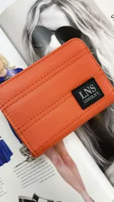 Billetera corta de diseño capitoneado con cierre perimetral y solapa frontal. Cuenta con compartimentos internos para tarjetas y billetes, e incluye una correa de mano desmontable.