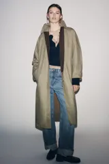 Trench coat de corte largo y diseño minimalista, confeccionado en tejido encerado color beige. Presenta cuello solapa, cierre frontal con botones oscuros y bolsillos laterales integrados.