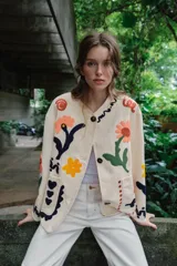 Chaqueta de algodón color crudo con cuello redondo, manga larga y bolsillos delanteros de plastrón. Presenta un diseño con bordados coloridos de flores y motivos abstractos a contraste, y cierre frontal con botones oscuros.
