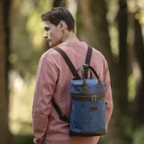 Mochila matera de diseño vertical, confeccionada en tela canvas azul con detalles en cuero sintético marrón. Cuenta con doble asa superior de mano, correas ajustables para la espalda, cierre metálico frontal y bolsillo trasero.