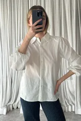 Camisa blanca de textura rayada vertical, con cuello clásico, cierre frontal de botones y mangas largas abullonadas con puños elásticos.