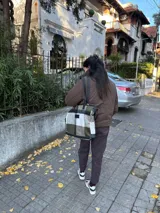 Bolso tipo tote de gran capacidad con diseño de cuadros en tonos verde oliva, blanco y negro. Presenta detalles en cuero negro en la base y en las correas de hombro, además de un parche de cuero con logo en el frente. Incluye cierre superior y bolsillo interior.