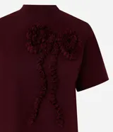Remera femenina de manga corta color bordó, con cuello redondo y una aplicación figurativa en forma de lazo en la parte delantera, hecha en tejido fruncido que crea relieve. Confeccionada en algodón certificado BCI.