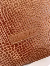 Mochila de cuero color marrón con textura tipo croco en el panel frontal. Cuenta con cierre superior, asa de mano superior, correas ajustables y una correa lateral extraíble con diseño estampado. Incluye logo de la marca grabado en el frente.