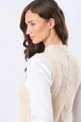 Chaleco tejido de punto grueso en color beige, con cuello redondo y diseño estructurado con detalles de trenzas verticales. Presenta un pequeño aplique de cuero sintético en la parte inferior.