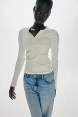 Remera ajustada de punto acanalado suave, color blanco, con cuello redondo ribeteado en picot y tapeta de botones. Es de manga larga.