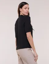 Blusa blanca de corte recto y tejido liviano de gasa con forro interno delantero. Presenta escote en V con cuello redondo y lazo central para anudar, mangas cortas con hilera de botones forrados en los laterales y ruedo ligeramente redondeado.