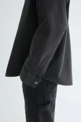 Sobrecamisa negra de lona gruesa de algodón, con cuello, tapeta con botones a presión y canesú en la espalda. Tiene mangas largas con botón a presión en los puños y dos bolsillos superiores de parche. El bajo es levemente redondeado y el corte es oversized.