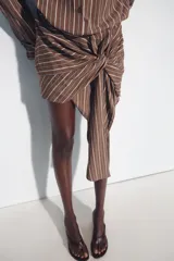 Vestido corto marrón con rayas verticales blancas, cuello camisero, manga larga y detalle de nudo y lazada en la falda. Cuenta con cierre frontal de botones.