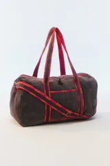 Bolso de viaje tipo duffel de color gris oscuro, confeccionado en algodón acolchado con pespuntes verticales. Cuenta con asas de mano y una correa larga regulable con detalles de tiras en tonos fucsia y naranja, cierre superior y herrajes metálicos dorados.