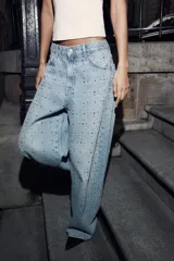 Pantalón de jean estilo baggy con tiro medio, corte holgado y diseño de cinco bolsillos. Presenta un acabado en lavado claro con aplicación de brillos distribuidos por toda la prenda.