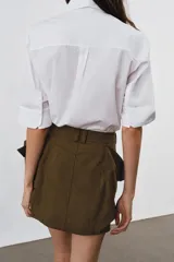 Camisa de algodón con cuello solapa y mangas por debajo del codo. Presenta un diseño con pechera plisada frontal, bajo recto y cierre mediante botones ocultos bajo solapa.