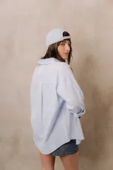 Camisa blanca de calce amplio, confeccionada en tejido liviano con sutiles rayas verticales, cuello clásico, mangas largas con hombros caídos y abotonadura frontal.