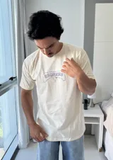 Remera de manga corta color beige jaspeado, con un estampado gráfico en la espalda que incluye el texto "TAS LOCO" y "SUNNY WITH A CHANCE OF STOKE" con un diseño de una persona en el agua con una ballena.